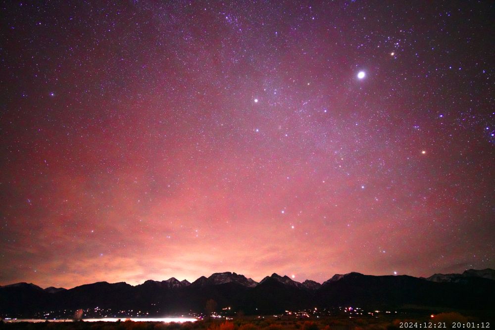 Bright red/purple airglow, with yellow air pollution on the horizon. Jupiter shines brightly top right near Taurus. The Rocky Mountains stretch across the foreground. There are houselights visible, and the moving light trail of a car. The sky is full of stars, the trail of Milky Way visible from center top angled toward bottom right. Mars is just coming up over the horizon.