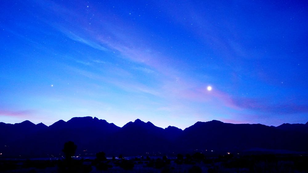 The sky is now a lighter shade of blue and overexposed on the horizon. Even fewer stars are visible. Venus is higher now over the silhouette of the Rocky Mountains, magnified by a thin layer of clouds that are turning a more brilliant pink. Pleiades is left and up a little from Venus. The lights of Crestone and Baca Grande homes sprinkle the foreground. Trees and chamisa in the foreground is now illuminated by dawn's first light.