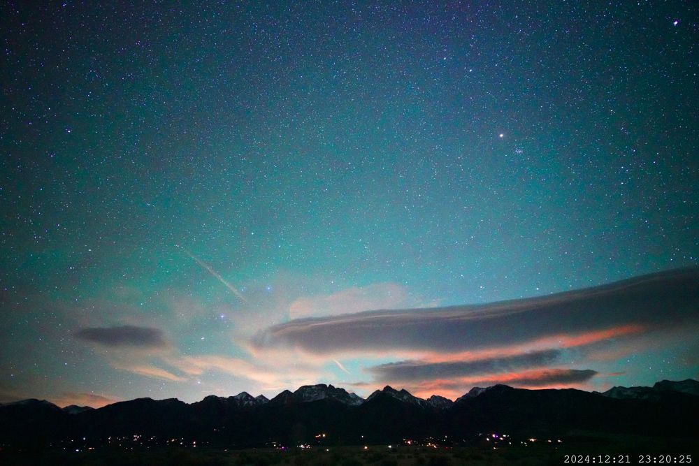 Bright green airglow fills the sky. There are lenticular clouds lit from underneath by the rising moon that is not yet in the frame. The clouds are fringed with reddish-orange light. The Rocky Mountains stretch across the foreground. There are houselights visible.