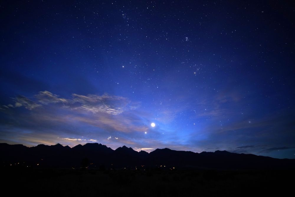 The silhouette of the mountains stretches across the lower quarter of the frame. Venus is a bright orb, magnified by wispy clouds over the mountains. Jupiter is a small orb lower left of Venus, very close. The clouds are tinged orange with first light. The Pleiades sit high above the scene. Orion lies on his side to the right, belt clearly visible and sword partly obscured by clouds. The sky is full of stars and ranges in color from light blue center around Venus, to dark blue at the top and in the corners. There are houselights visible in the foothills