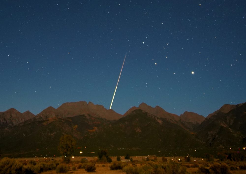 October 3, 2025 at 1:57am MDT

Crop of previous frame. A meteor streaks downward in the center of the frame, disappearing behind the ridgeline of the Rocky Mountains. There are stars visible in the sky which is tinged blue, illuminated by the waxing gibbous moon. The meteor has a fainter pink/purple tail that abruptly shifts brighter, halfway through the streak. The foreground is mostly brown grasses and chamisa.