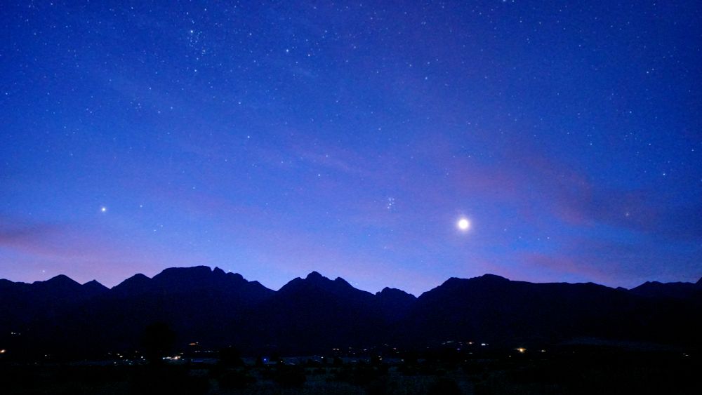 The sky is lightening up and appears a beautiful shade of blue. Many stars are still visible. Venus is higher now over the silhouette of the Rocky Mountains, magnified by a thin layer of clouds that are turning a nice shade of orange/pink. Pleiades is left and up a little from Venus. The lights of Crestone and Baca Grande homes sprinkle the foreground.
