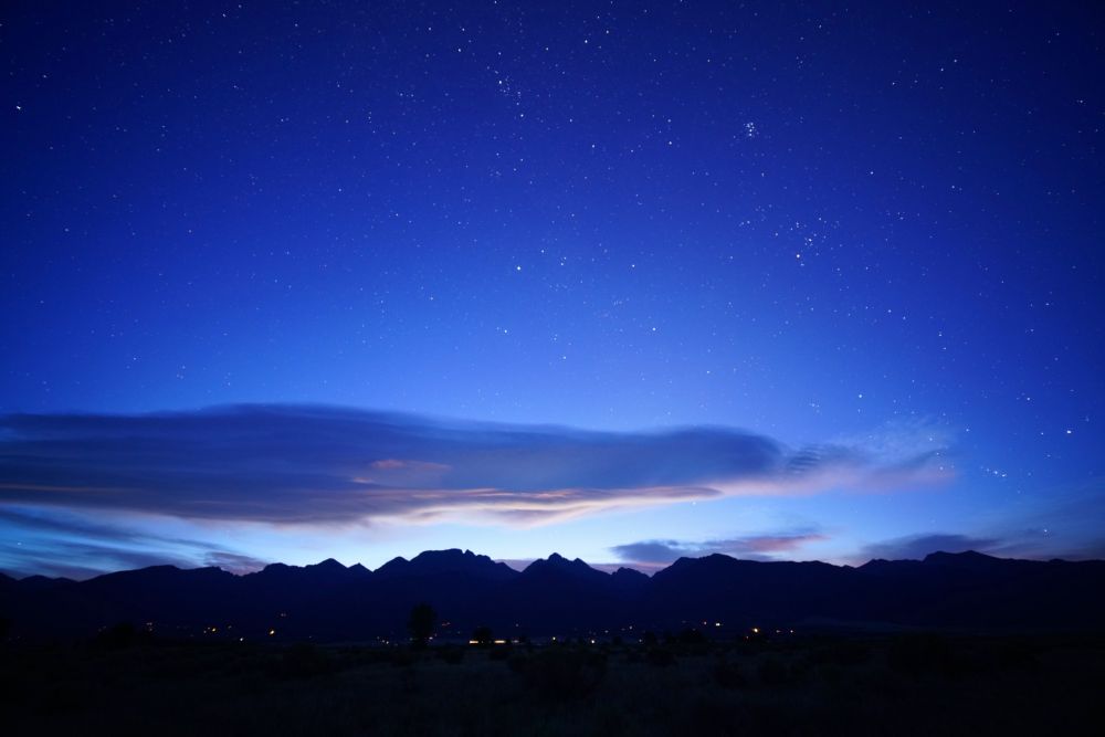 The sky is much lighter and fewer stars are visible, though the Pleiades and Orion are still distinct. A long lenticular cloud hangs over the Rocky Mountains  The sky is lighter blue now. Lights from homes are still visible in the foothills of the mountains. 