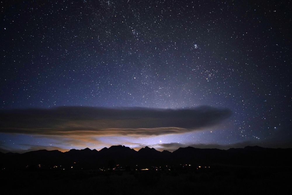 The sky is full of stars. A long lenticular cloud hangs over the Rocky Mountains. Light pollution from the Front Range illuminates the underside yellow. The sky is purple to black. Lights from homes are visible in the foothills of the mountains. The Pleiades site right of center.