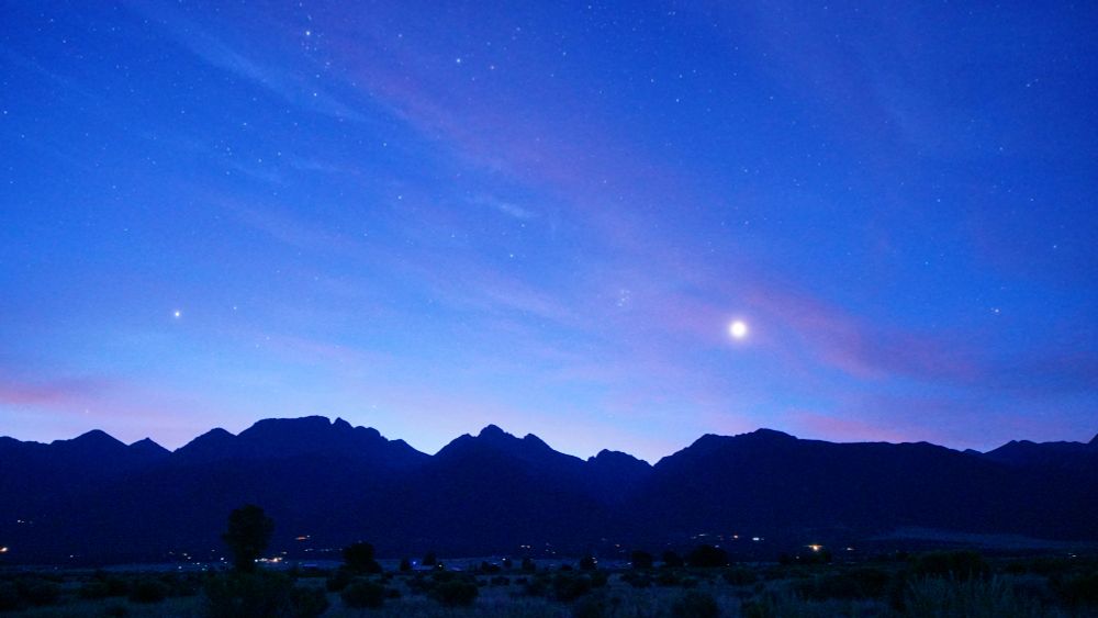 The sky is now a lighter shade of blue. Fewer stars are visible. Venus is higher now over the silhouette of the Rocky Mountains, magnified by a thin layer of clouds that are turning a more brilliant pink. Pleiades is left and up a little from Venus. The lights of Crestone and Baca Grande homes sprinkle the foreground. Trees and chamisa in the foreground is now illuminated by dawn's first light.