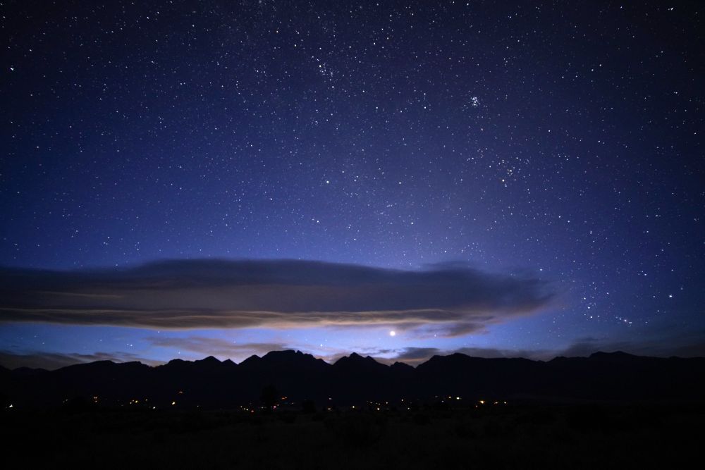 The sky is full of stars and lighter than the last frame. A long lenticular cloud hangs over the Rocky Mountains with Venus just below it. Light pollution from the Front Range illuminates the underside yellow. The sky is purple to black. Lights from homes are visible in the foothills of the mountains. The Pleiades site right of center. Orion is rising and the belt and sword are visible on the right.