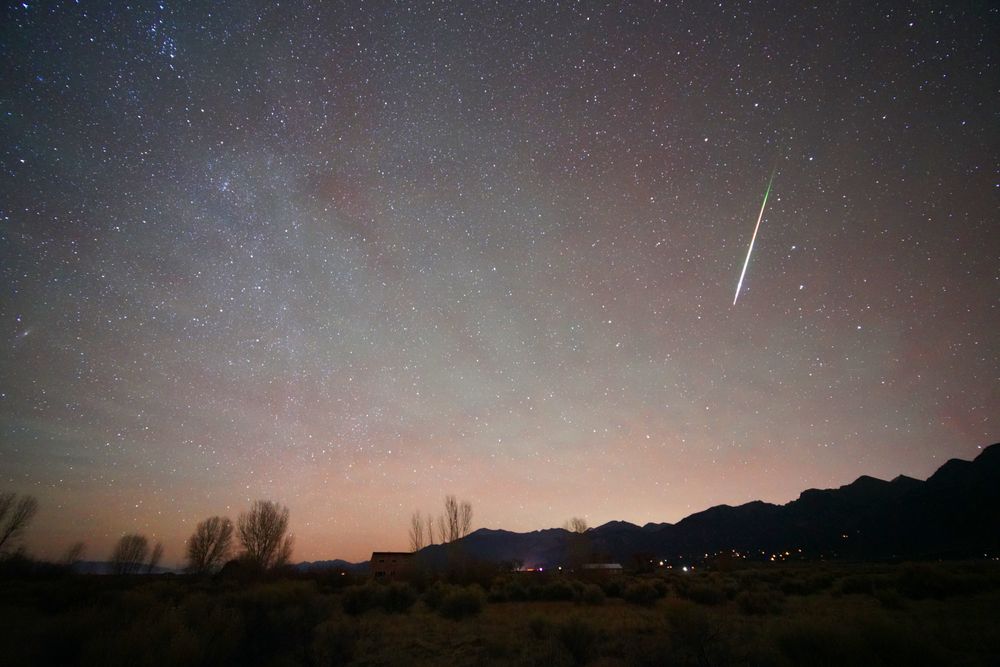 2025:11:16 03:32:07 MDT

A meteor streaks down on the right third of the frame. The tail of the meteor at the top is green. The body is white. The sky has patches of red and green airglow that are rippled with gravity waves. The Milky Way points down on the left. The sky is dark and full of stars. The Rocky Mountains stretch up along the horizon center to right. Houselights of Crestone and the Baca Grande POA dot the foothills. The bare branches of cottonwood and aspen trees stick up in the foreground (left and center)