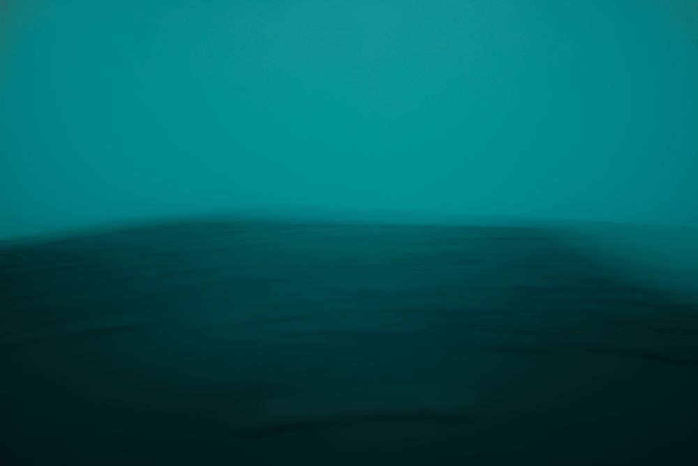 A dark aqua landscape barren with small bumpy patterns running across it. Misty teal hovers above and over the landscape in the background.