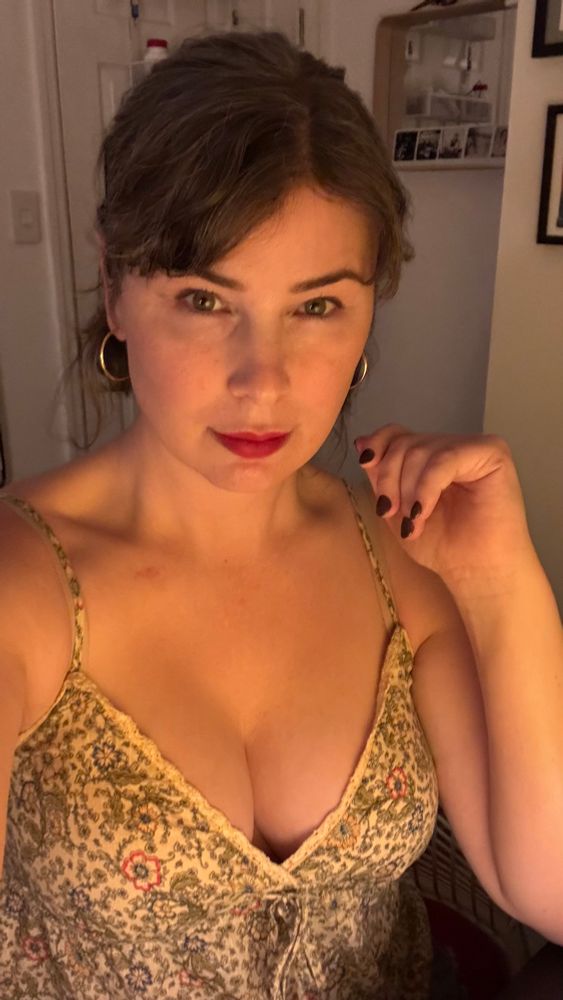selfie of a white woman with pulled back brown hair and red lipstick. she’s wearing a floral tank top that displays cleavage. her hand is held up and she has burgundy nail polish