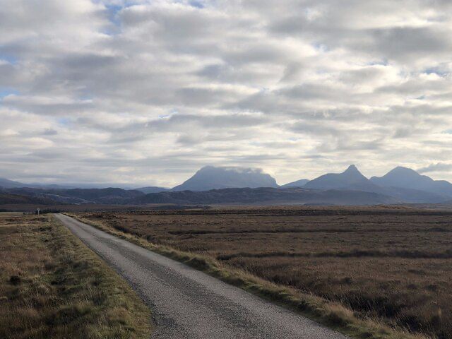 A road crosses a vast bog. There are lines in the vegetation where peat had been dug. There is a patchy cloudy sky and isolated hills in the distance. The one on the left, Cùl Mòr has its summit in the cloud. To the right a sharp peak, Stac Pollaidh and another simple triangular peak Cùl Beag are clear of cloud.
