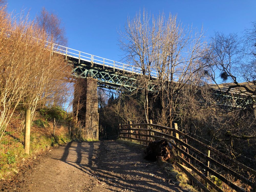 Gravel road passing under a high railway bridge. The viaduct is made of green girders supported by stone pillars. On the right of the road there is a steep descent into a gorge so the track is high above the burn. It is a bright sunny day with a blue sky. Trees bare at the start of Winter.