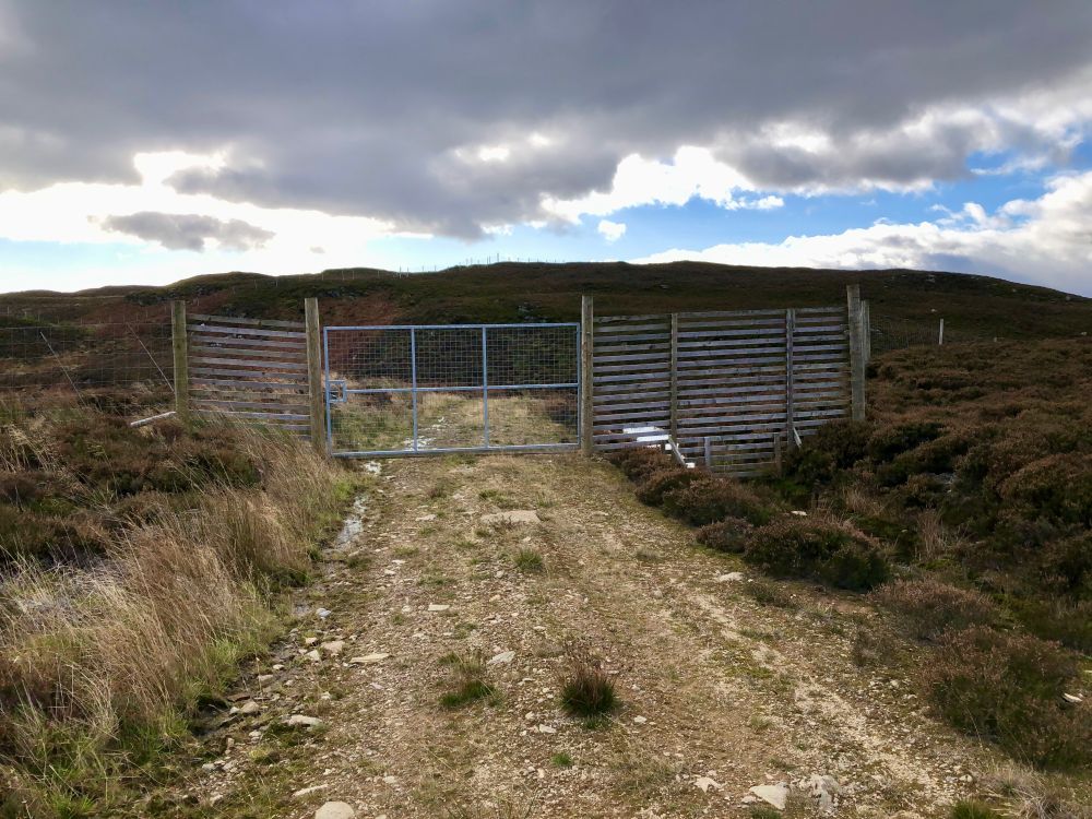 Roughly bulldozed track on heather moorland. There is a tall fence and mesh gate in the middle distance. The wood beside the gate is poor quality and soon will not tolerate anyone climbing it. A hazard.