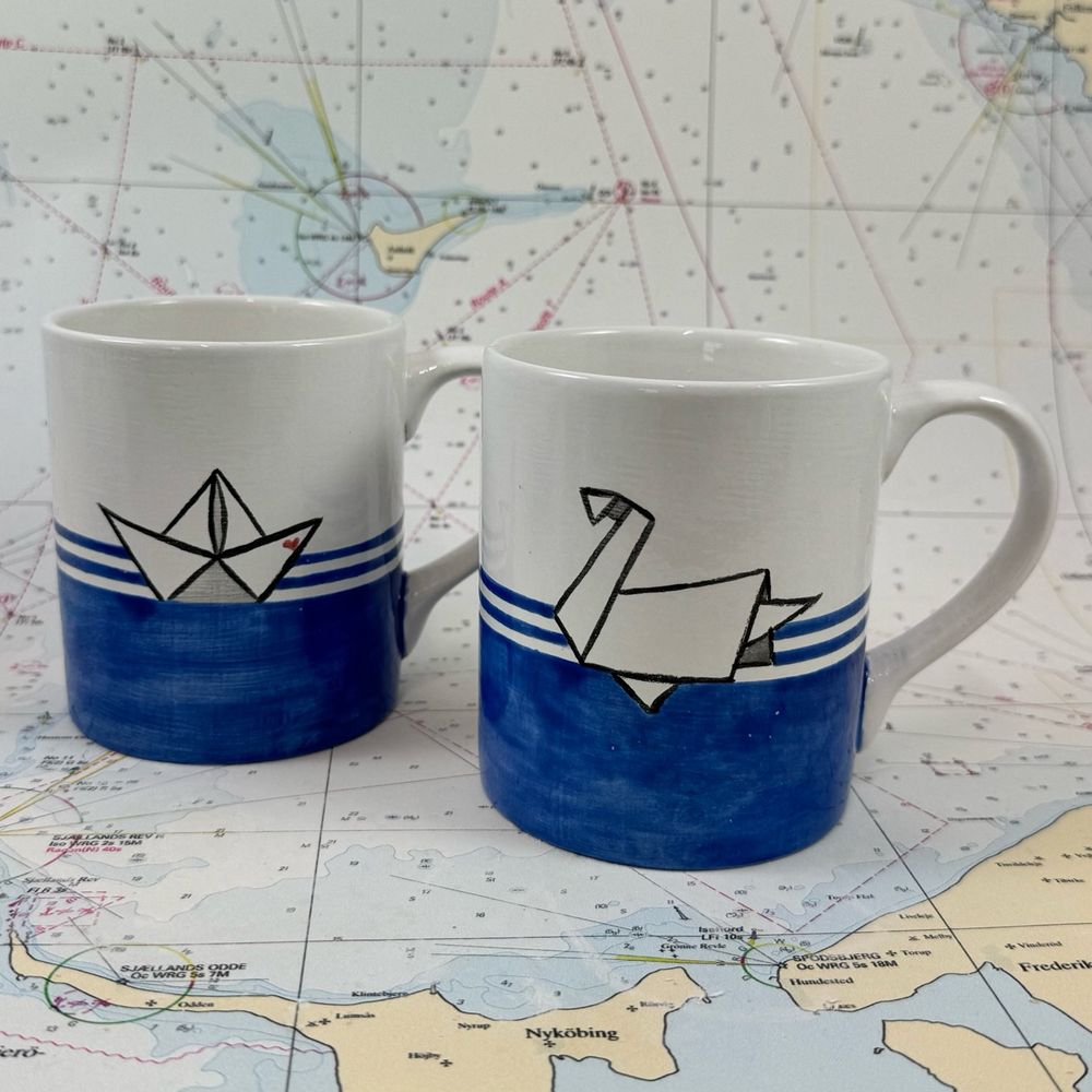 Henkelbecher aus Keramik. Handbemalt mit blau weißen Streifen und ein Becher mit Origami-Schiffchen der andere mit einem Origami-Schwan. Fotografiert vor einer alten Seekarte.