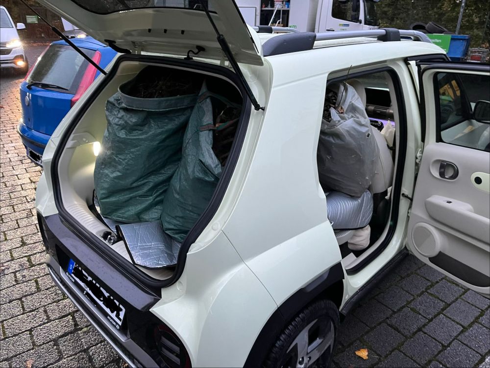 Foto (von schräg hinten rechts aufgenommen) eines auf einem Recyclinghof parkenden cremefarben lackierten Hyundai Inster mit geöffneter Heckklappe und geöffneter rechter hinterer Seitentür. Im Inneren des Autos sieht man zwei große grüne Gartenabfallbehälter und einen kleineren transparenten Gartenabfallsack. Nur erahnen kann man, dass dahinter noch vier weitere transparente Abfallsäcke im Auto verborgen sind.
Das Foto vermittelt einen Eindruck davon, wieviel Stauraum in diesem Kleinwagen steckt, der nur 3,83 Meter lang und 1,61 Meter breit ist (Höhe 1,58 Meter).