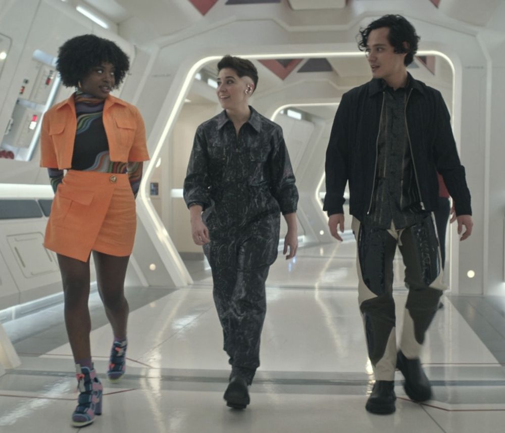 Screencap from Strange New Worlds season 3 episode 2. Three characters walk down a high tech white hallway, one is wearing a matching orange skirt and jacket combo with a black bodycon top under it that has waves of colourful stripes along with some very funky heel boots that seem to be fastened with bright pink straps. The middle person is wearing a dark patterned jumpsuit made of a satin type material swith a large collar, they've rolled thier sleeves rolled up. The third person is wearing a fairly conventional button up shirt and casual jacket, but the pattern on their pants is what's interesting. I don't even know how to describe it, are they chaps?? Hard to tell. They're funky, that's for sure