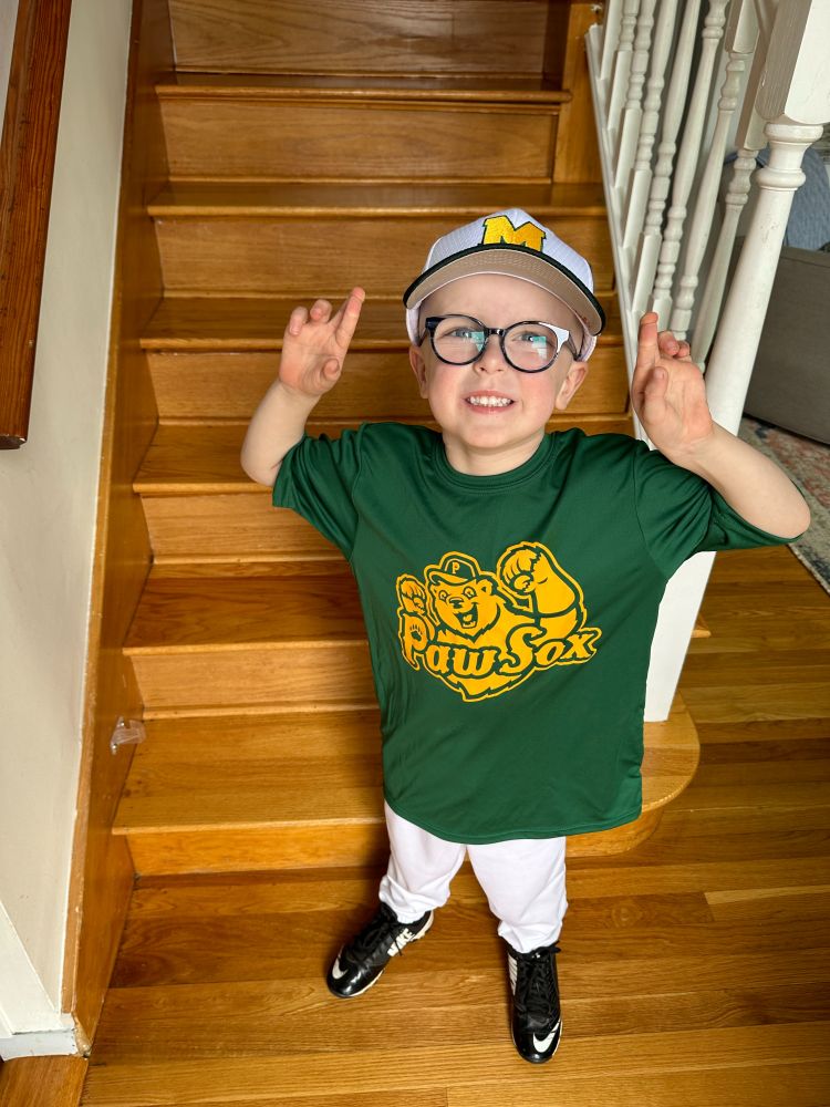 Owen smiles with his hands up, wearing a green shirt and white pants. He is standing in front of a staircase. 