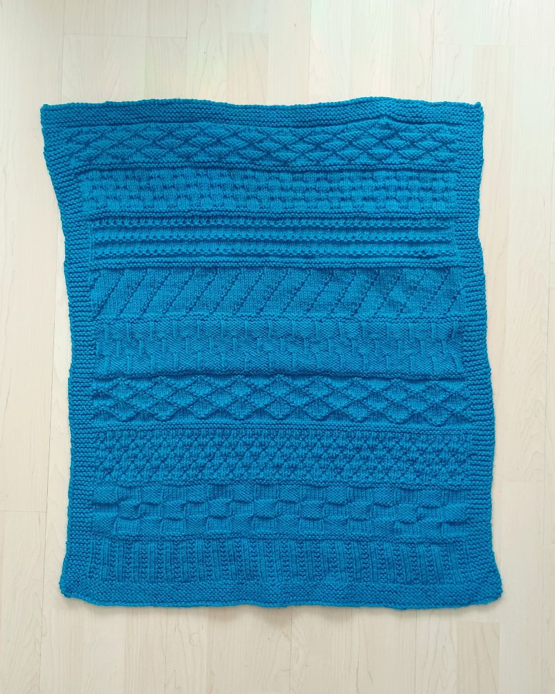 A blue, handknit baby blanket featuring stripes of different texture patterns and a garter stitch border. 