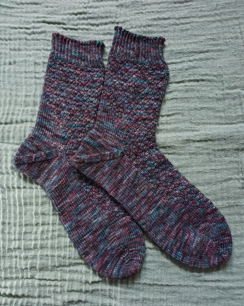 A pair of handknit socks made with dark, variegated yarn in cool tones. There is a textured diamond motif across the top and leg off the sock. 