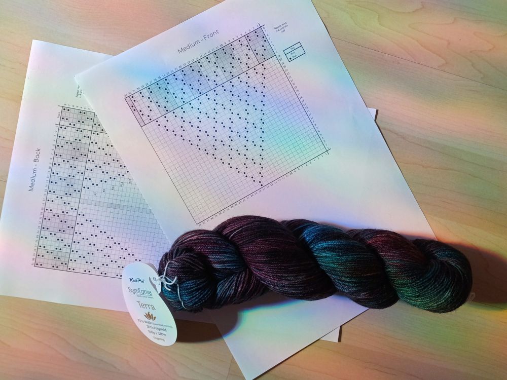 A skein of dark, variegated yarn in blue and purple tones lays on top of printed out knitting charts. The charts show a textured diamond motif. 