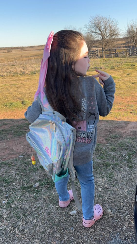 A 6 year old girlie with a big pink bow in her brunette hair and a holographic backpack slung over one shoulder. She’s throwing a peace sign over her shoulder and doing a kissy face. She has a school pride grey sweatshirt on, jeans, and sparkle crocs. She is standing outside with a rustic corral in the background and farm land. 