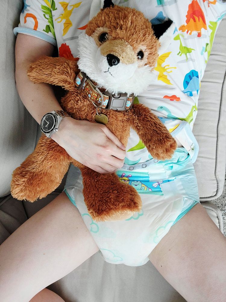 Me laying on the couch, cuddling my plush fox. I'm wearing a colorful onesie with a dinosaur print and a soaked Kiddos diaper underneath.