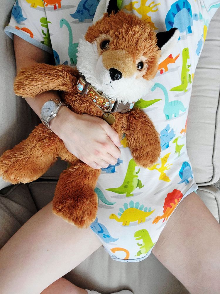 Me laying on the couch, cuddling my plush fox. I'm wearing a colorful dinosaur print onesie and a soaked TryAgain diaper underneath.