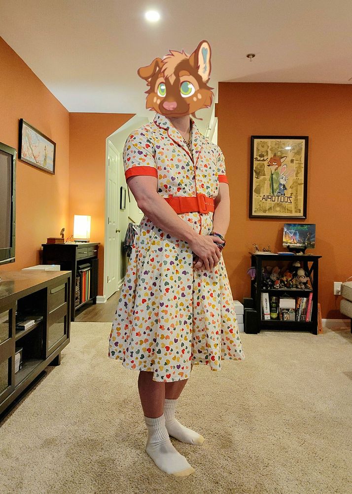 Me standing in my living room, hands clasped in front of me. I'm wearing a 1950s style white shirt dress, trimmed in bright red, with colorful hearts all over. It has a bright red belt to match.