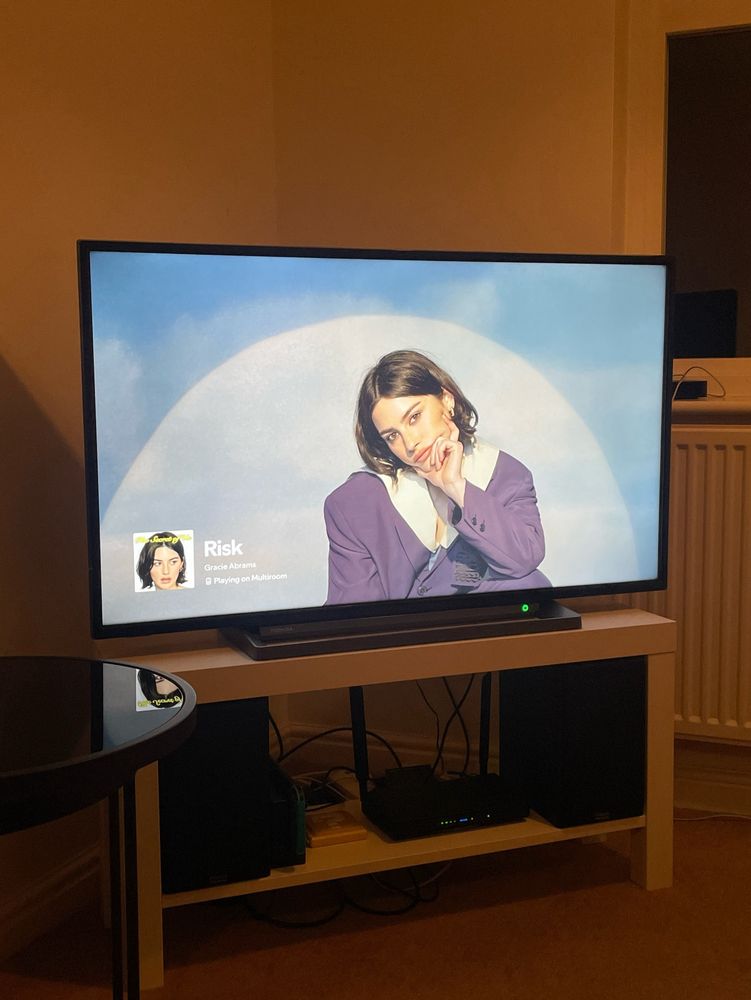 a tv stand displaying Spotify playing Risk by Gracie Abrams and two black speakers below 