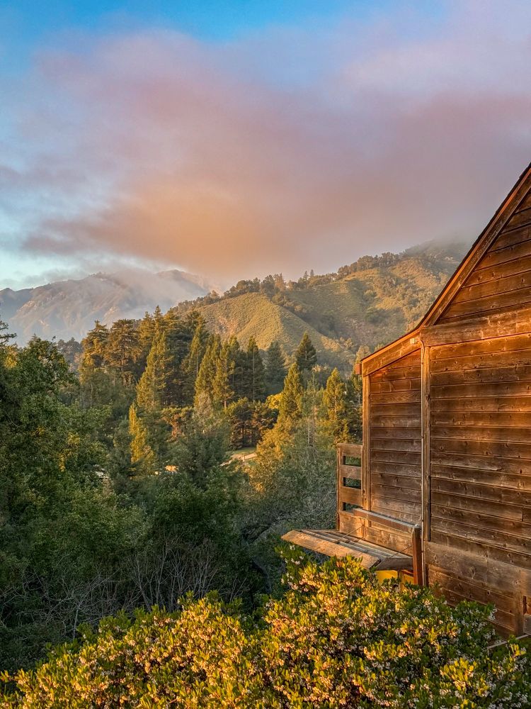 A photo of mountains covered in trees. There’s a cabin in the foreground overlooking a valley. There are distant mountains and low hanging clouds. It’s “golden hour” in the photo and there’s a warm hue. 