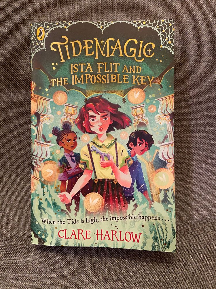 The book cover for Tide Magic: Ista Flit and the Impossible Key by Clare Harlow has a green background and illustrations of a red haired girl with red bobbed hair wearing a iight green shirt and red trousers holding a large grey key. Behind her are a black girl in a purple shirt with her hair in bunches and an Asian boy in a blue shirt with unruly hair. The title is at the top and the author's name below.