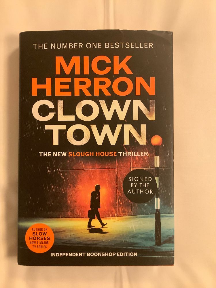 The book cover for Clown Town by Mick Herron shows the author’s name in capital letters in orange at the top underneath the words The Number One Bestseller and the title underneath the author’s name in white block capitals. Underneath the title is the subhead The New Slough House thriller. There’s an image of a male figure carrying a briefcase walking on a wet pavement. It’s raining and there’s the amber streetlamp showing a pedestrian crossing. There is a round black sticker saying it has been signed by the author and an orange sticker saying that Slow Horses is now a major TV series. At the bottom are the words Independent Bookshop Edition.