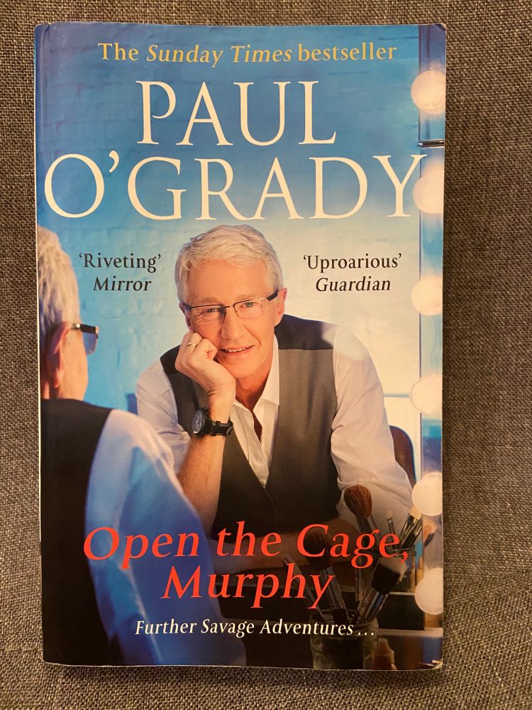 Book cove for Open The Cage, Murphy by Paul O'Grady shows Paul O'Grady looking into a mirror and back out at the reader. He is wearing glasses and a brown waistcoat over a white shirt and a watch with a black strap. His name is at the top of the cover and the book title is in red at the bottom of the cover