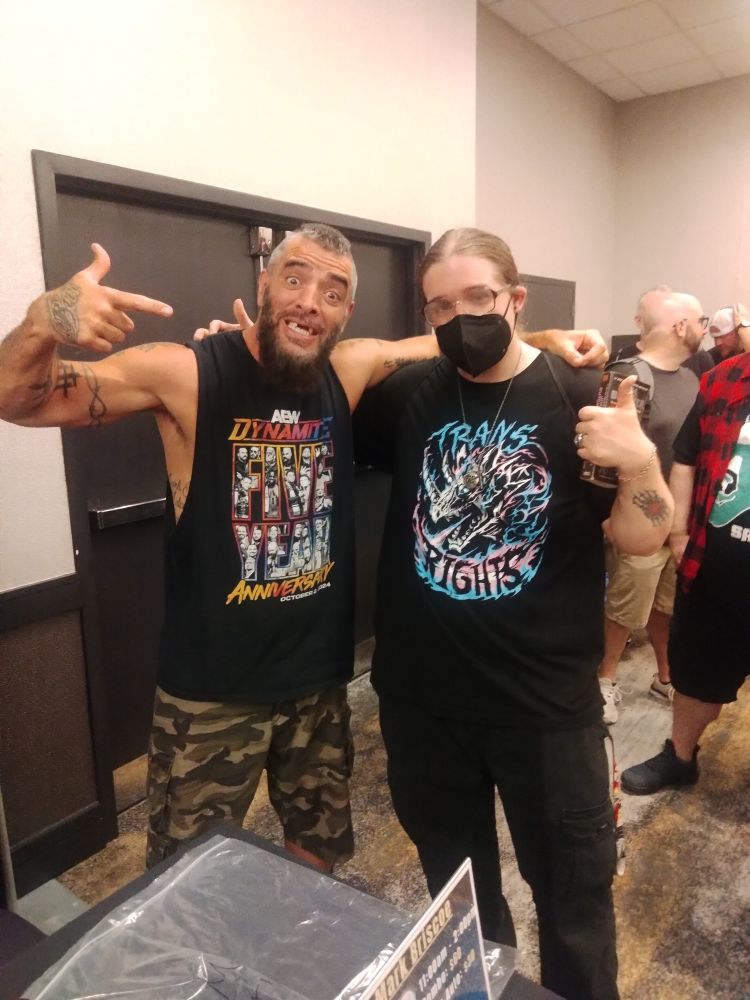 Wolgs and Mark Briscoe with an arm across each other's shoulders, wolgs holding a book with a thumbs up, Briscoe pointing over at him. Briscoe is wearing camo shorts and a sleeveless shirt celebrating the fifth anniversary of AEW Dynamite  