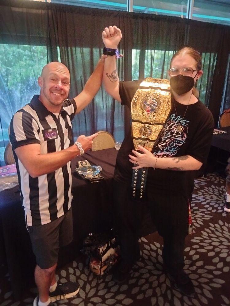 Bryce Remsburg in shorts and a referee shirt with a patch saying "All In Texas" pointing at wolgs and holding his arm up as if wolgs had just won a championship. Wolgs is holding a copy of the AEW international championship over one shoulder and holding it steady with the other hand.  