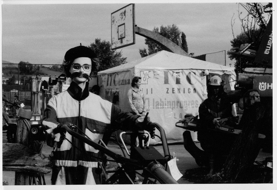 Female mannequin with pasted on mustache, glasses and black beret, dressed in a warm-up suit, posed as if operating a rototiller in a trade-fair display of outdoor farm and garden equipment. Behind her is a tent with wording "ITC Zenica labinprogres" and behind that is a concrete basketball backboard with the hoop missing. Overall, a jumble. There's a live woman standing in front of the tent, smiling at someone or something to her left.