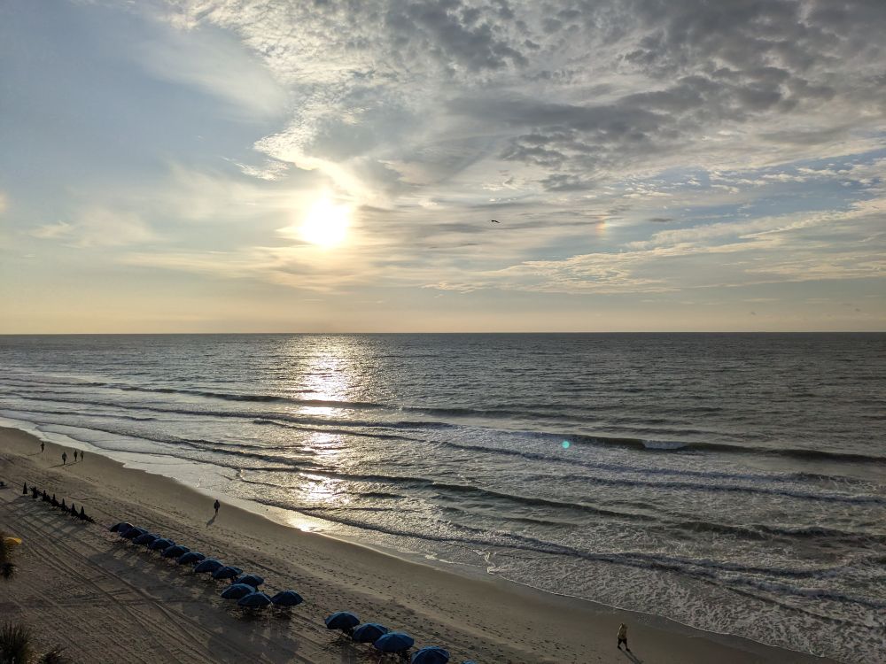 A view of the beach and ocean from an elevated position. The Sun is low in the sky but rising and partially hidden by light cloud cover with its light reflecting off of the water.