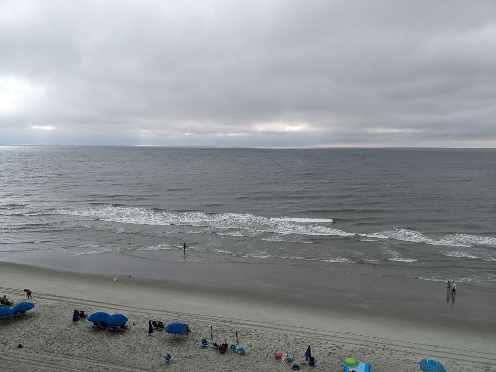 An elevated view of the beach and ocean with a heavily overcast sky and distant rain on the horizon.