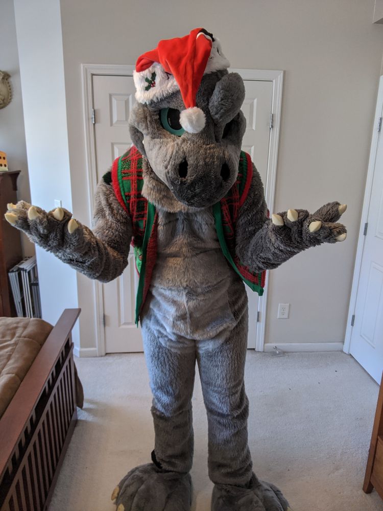 Nullstrukt the dimetrodon fursuit shrugging while wearing an ugly Christmas sweater vest (with T-Rexes on it) and a Santa hat.