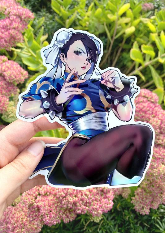 A sticker of Chun Li from street fighter being held in front of a bush with pink flowers. Chun Li is holding up her fists in an action pose. 