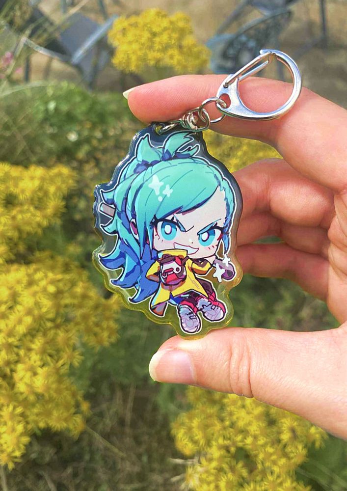 A hand holding a chibi style illustration charm of Mizuki from AI: The Somnium Files. She grinning mischievously holding her metal pipe. The picture has been taken outside in front of some yellow flowers.