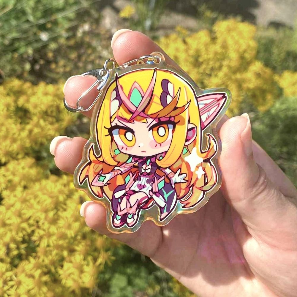A hand holding a chibi style illustration charm of Mythra from Xenoblade chronicles 2. She is holding her sword in a battle pose and frowning (but it's cute). The art is adorned with sparkles. The picture as been taken outside in front of some yellow flowers.