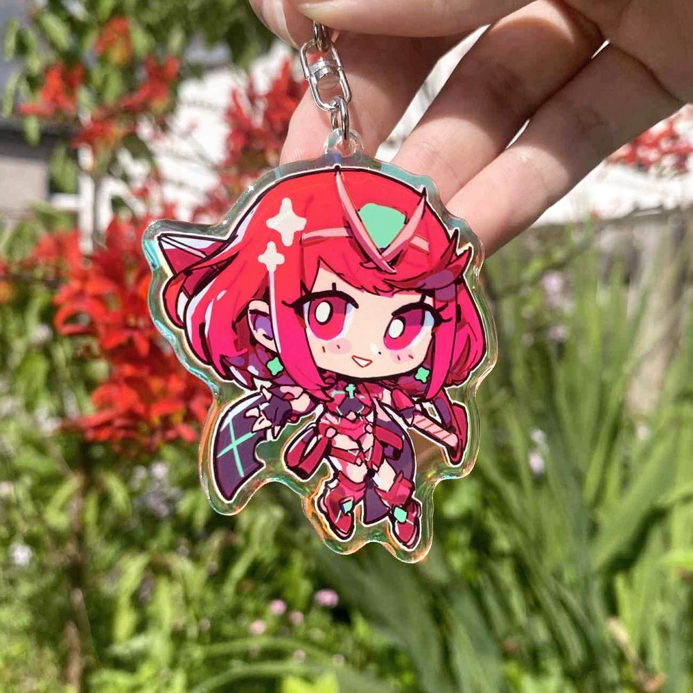 A hand holding a chibi style illustration charm of Pyra from Xenoblade chronicles 2. She is holding her sword in a battle pose and smiling. The art is adorned with sparkles. The picture as been taken outside in front of some red flowers.