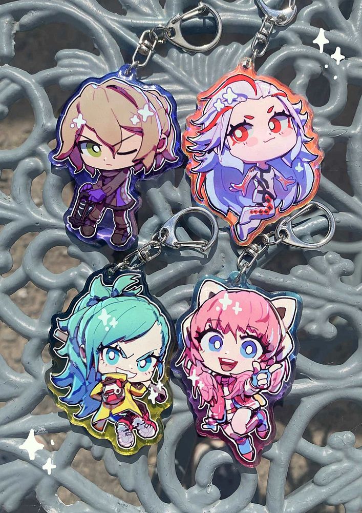 Four charms of chibi style illustrations characters from AI: The Somnium Files (Date, Aiba, Mizuki, Iris) laid out on an outdoor table. The image is adorned with sparkles.