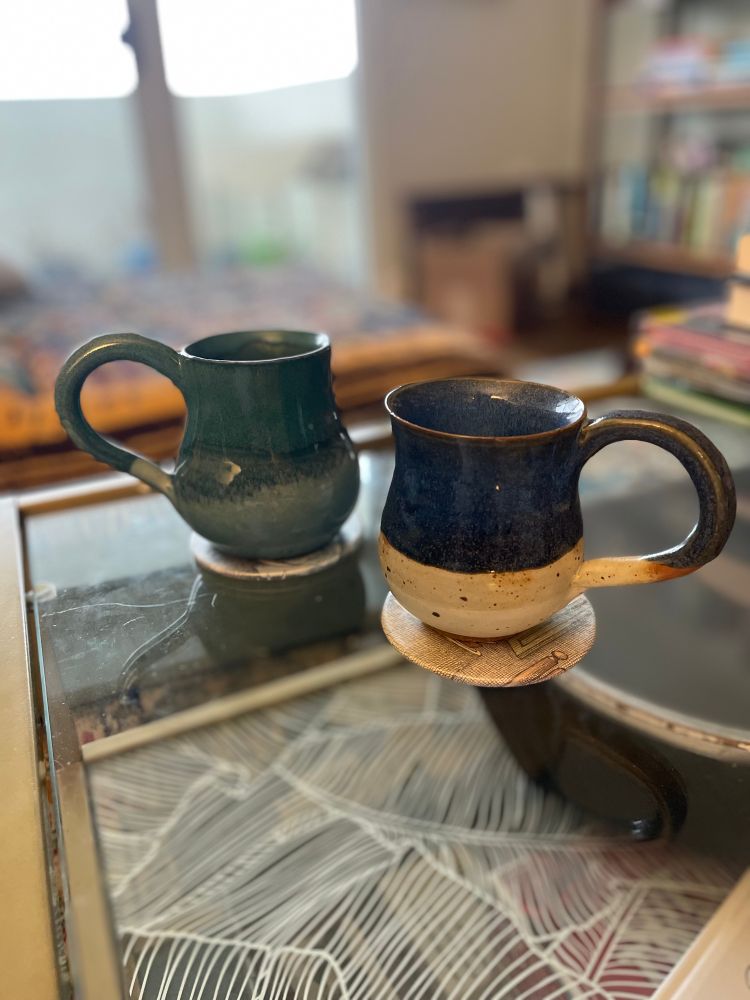 Photo of two mugs of tea on a glass coffee table. The mug on the left is shades of teal, with a large, swooping handle. The mug on the right has a grey stone base, with dark blue glaze on top. The handle is slim and elegant. Bright winter light filters in through a window in the background.
