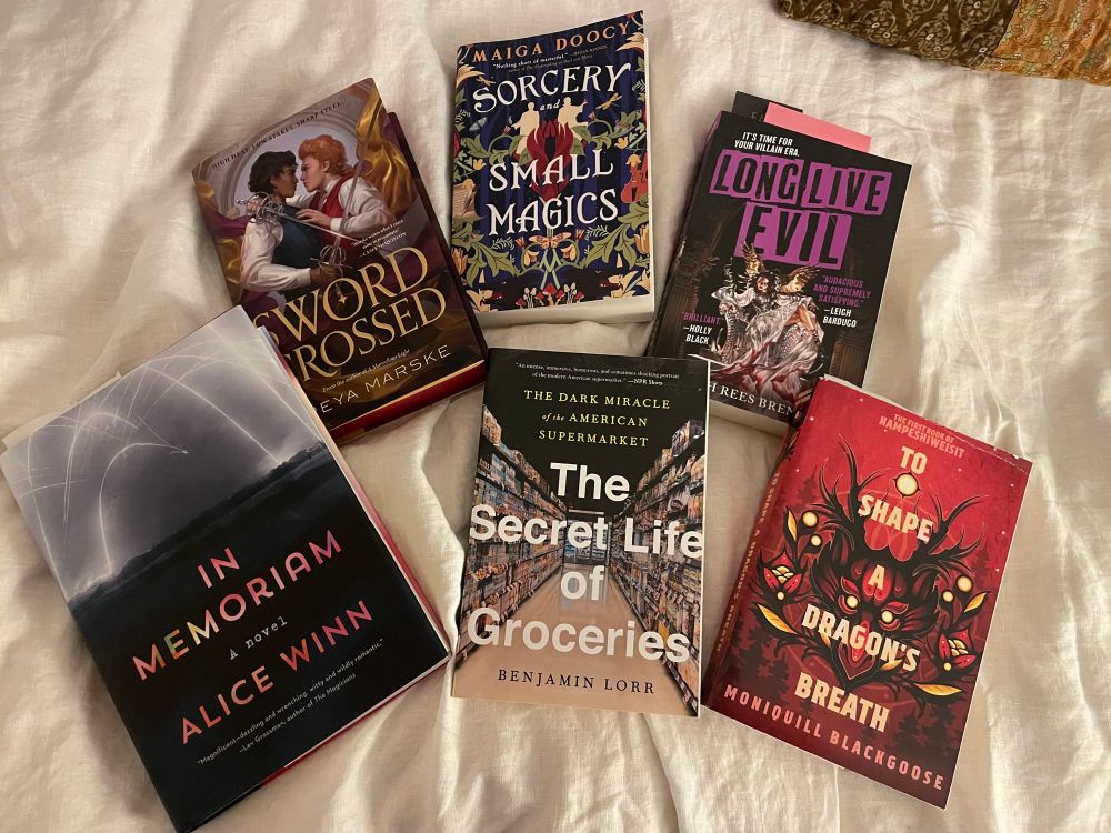 Photo of 5 books spread across a white bedspread. From left to right, the books are:
Swordcrossed by Freya Marske
Sorcery and Small Magics by Maiga Doocy 
Long Live Evil by Sarah Rees Brennan
In Memoriam by Alice Winn
The Secret Life of Groceries by Benjamin Lorr
To Shape a Dragon’s Breath by Moniquill Blackgoose