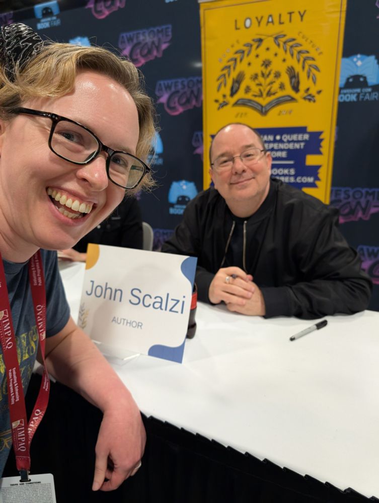 A photo of a woman in a blue t-shirt smiling like a dork because she has just met one of her favorite authors, John Scalzi. He is also in the photo and is being very tolerant of the photo op.