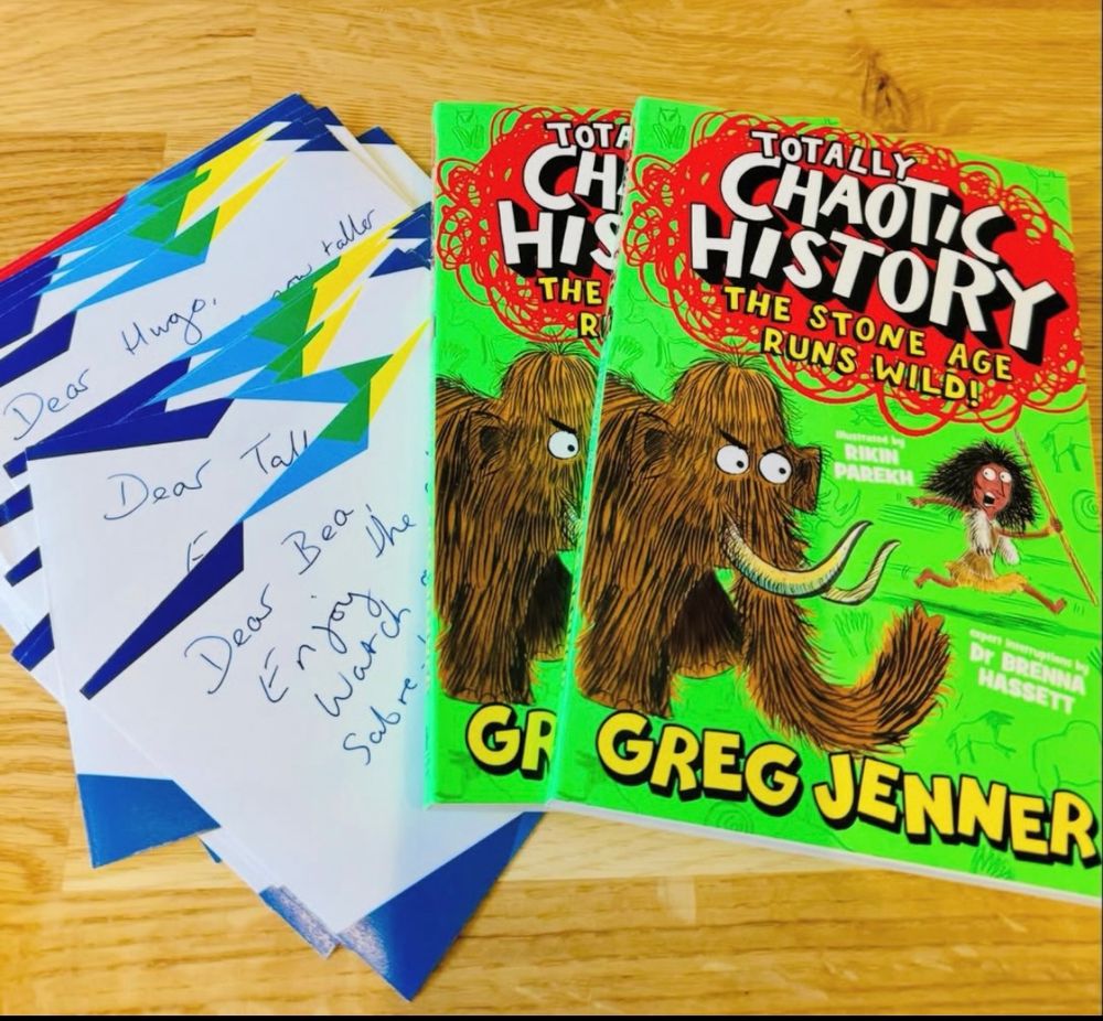 The cover of Greg Jenner & Dr Brenna Hassett’s book TOTALLY CHAOTIC HISTORY: THE STONE AGE RUNS WILD has a line green cover and a hilarious cover showing a woolly mammoth chasing a Stone Age hunter 

Next to it is a pile of personalised stationery showing Greg’s handwritten messages to kids which get stuck in the front of the book 