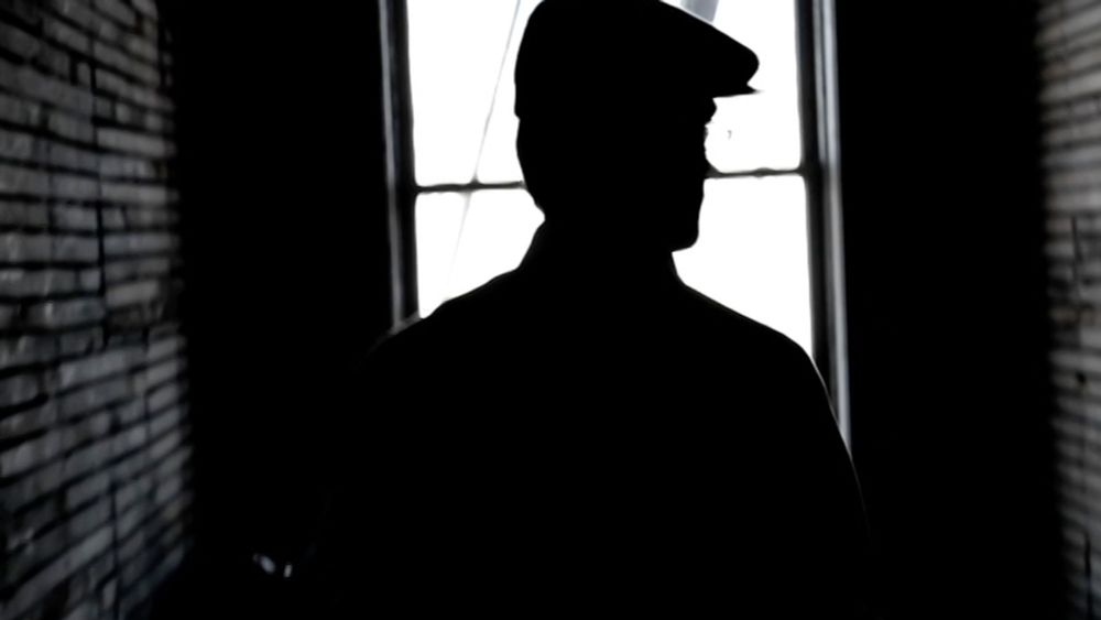 Shadowed profile of a man in a flat cap — standing inside a dimly lit space, outlined against a bright window, with rough, exposed wall studs running vertically along the left side.