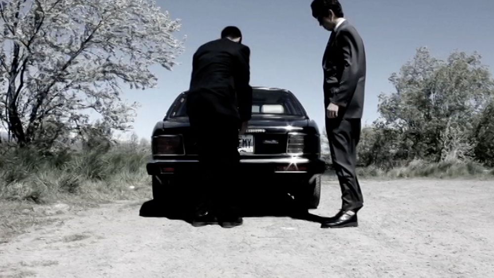 Two men in black suits stand behind a black car on a dirt road — one bends toward the trunk, the other stands upright watching, with bright daylight and a leafless tree in the background.