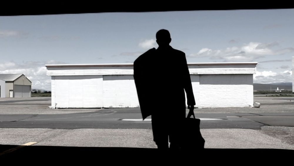 Silhouette of a man in a suit walking toward an empty airfield — carrying a briefcase, framed against a bright sky and white hangar buildings in the distance.